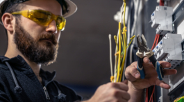 A male electrician works in a switchboard with an electrical connecting cable, connects the equipment with tools.