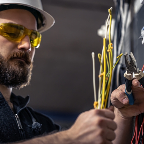 A male electrician works in a switchboard with an electrical connecting cable, connects the equipment with tools.