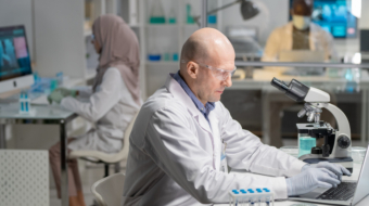 Mature serious male lab worker in gloves, whitecoat and protective eyeglasses searching for scientific information in the net